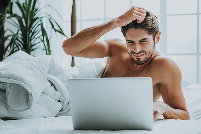 Mann liegt im Bett fasst sich in die Haare und schaut auf sein Laptop