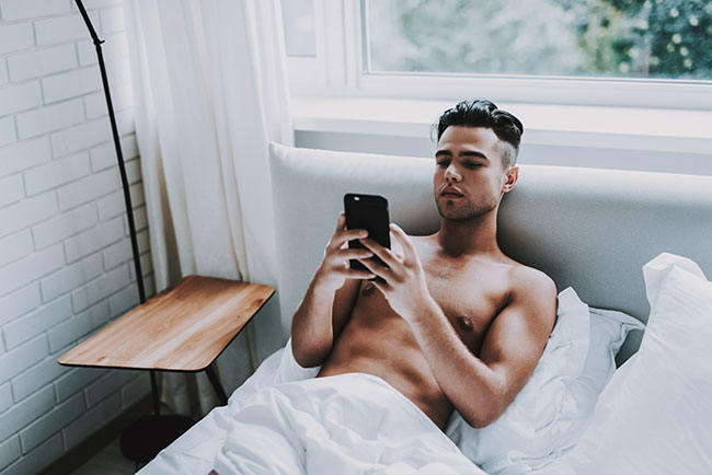Mann liegt oberkörperfrei im Bett und schaut auf sein Smartphone
